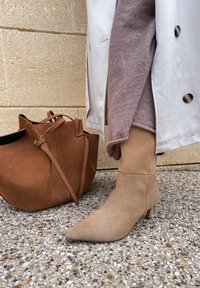 Botine din suede bej, cu vârful ascutit și toc de 5 cm, asortate cu o geantă tote din suede maro, pe un fundal texturat gri.