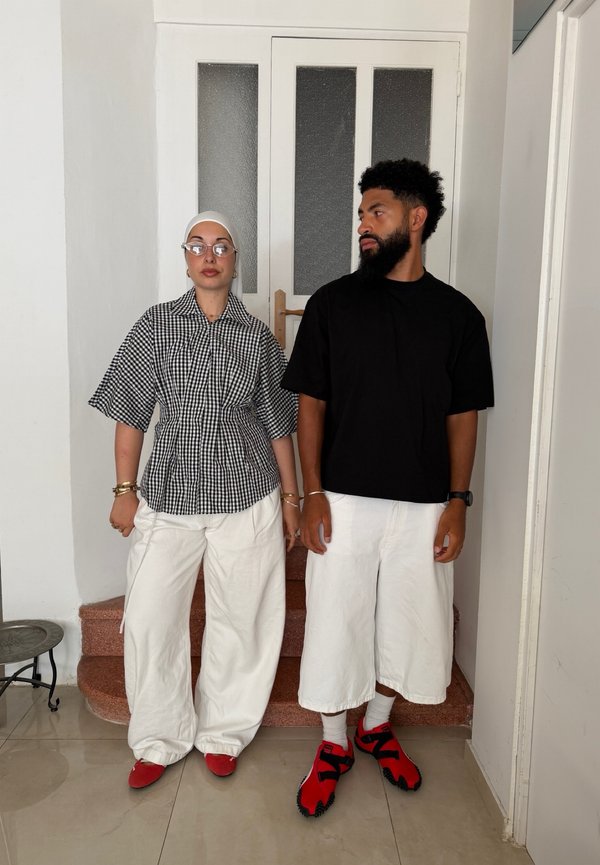 Due persone sono all’interno vicino a una porta, indossano pantaloni bianchi e scarpe rosse; una indossa una camicia nera, l’altra una maglietta a quadri bianca e nera.