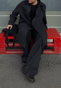 Άτομο ντυμένο με μαύρο παλτό και φαρδιά παντελόνια, καθισμένο σε κόκκινο ξύλινο παγκάκι, κρατώντας με το ένα χέρι μια μαύρη τσάντα.