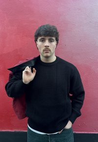 Giovane uomo con baffi che indossa un maglione nero e jeans, con una giacca rossa appoggiata sulla spalla, in piedi di fronte a una parete rosa con texture.