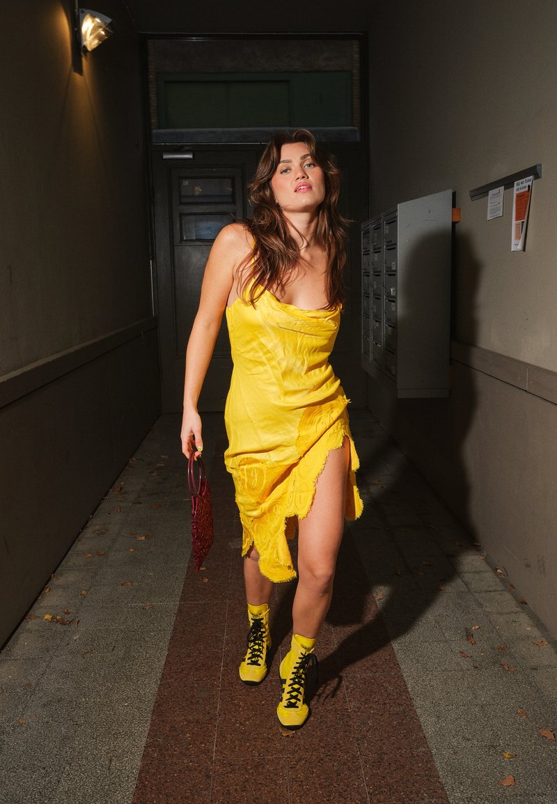 Robe en satin jaune avec bordure en dentelle, fente ouverte, assortie à des baskets montantes noires et jaunes et un sac à main rouge à perles. Cadre intérieur.