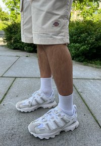 Sneakers atletici bianchi con accenti grigi, caratterizzati da una parte superiore in rete, suola testurizzata e design con lacci. Indossati con pantaloni corti beige e calze bianche.