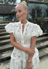 Γυναίκα με ξανθά μαλλιά μαζεμένα σε κότσο, φορώντας ένα λευκό δαντελένιο φόρεμα, χρυσά κοσμήματα και κόκκινο βερνίκι νυχιών, στέκεται κοντά σε πέτρινες σκάλες σε εξωτερικό χώρο.