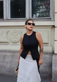 Femeie îmbrăcată într-un top negru fără mâneci și o fustă albă, cu ochelari de soare închiși la culoare, purtând o geantă împletită, stând în fața unui perete texturat și a unei feronerie.