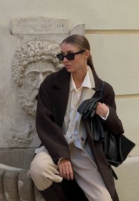 Černý oversized kabát, bílá blůza, béžové kalhoty, černé kožené rukavice a černá taška. Pozadí zahrnuje kamennou zeď a vyřezávanou tvář.