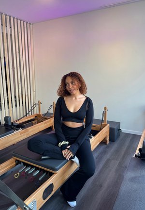 Mujer vestida con ropa deportiva negra sentada con las piernas cruzadas sobre una máquina de Pilates reformer en un estudio luminoso con suelo de madera clara y divisor de ambiente.