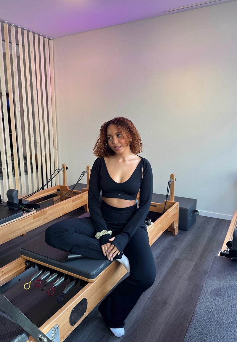 Femme en tenue de sport noire assise en tailleur sur un appareil de Pilates dans un studio lumineux avec un sol en bois clair et un séparateur de pièce.