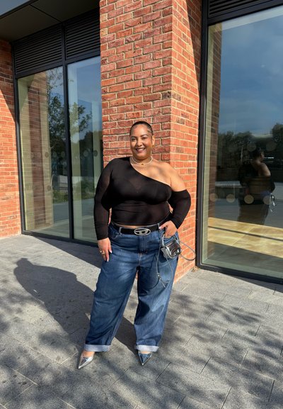 Top negro de un solo hombro y manga larga combinado con jeans anchos azules, tacones puntiagudos plateados y un pequeño bolso negro. Pared de ladrillo en el fondo.