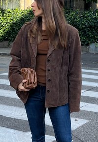 Brown suede jacket with notched lapels and black buttons, worn over a brown top and blue jeans. Holding a woven brown handbag.