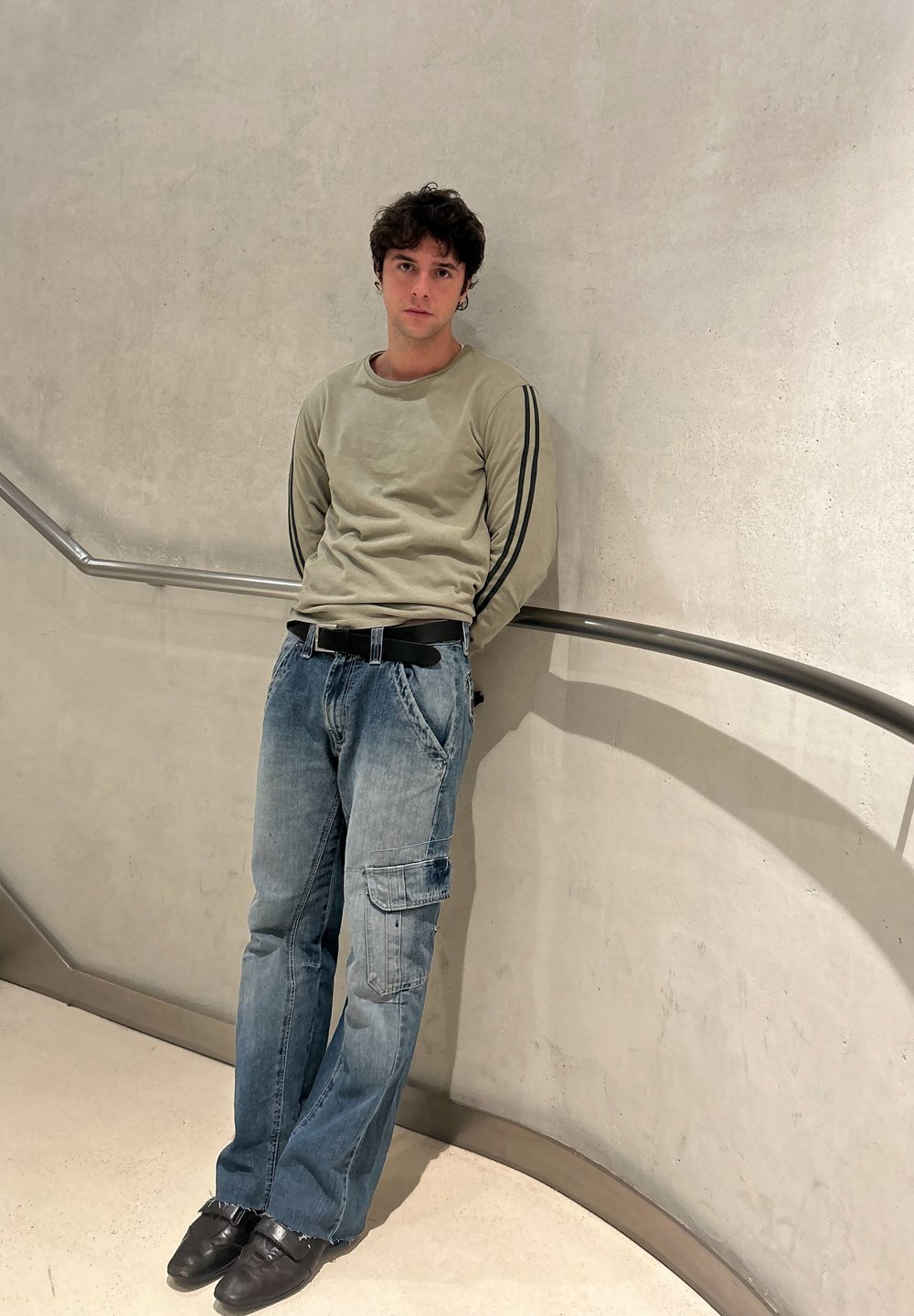 Jeune homme aux cheveux bouclés se tient contre un mur incurvé avec une rampe métallique, portant un sweat-shirt vert clair, un jean bleu et des chaussures noires.