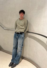 Jeune homme aux cheveux bouclés se tient contre un mur incurvé avec une rampe métallique, portant un sweat-shirt vert clair, un jean bleu et des chaussures noires.