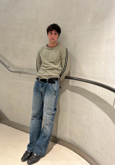 Joven con cabello rizado que se apoya contra una pared curva con pasamanos metálico, lleva una sudadera verde claro, jeans azules y zapatos negros.