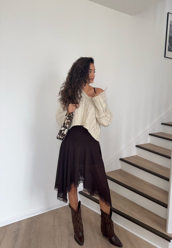 Vrouw met krullend donker haar draagt een crèmekleurige kabelgebreide trui, asymmetrische zwarte rok, bruine laarzen met studs en houdt een tas met luipaardprint vast bij een trap.