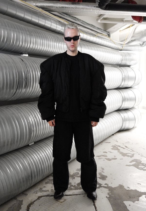 Persoon met kort blond haar en zwarte zonnebril, draagt een oversized zwart jack en broek en staat in een industriële ruimte met metalen kanalen.