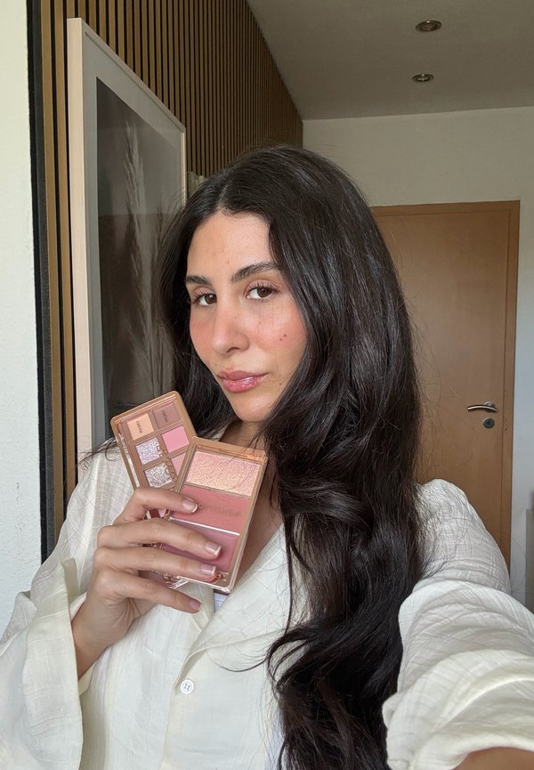 Kvinde med langt mørkt hår holder to makeup-paletter, der viser nuancer af pink og neutrale farver inde i et rum med en trædør og kunstværk.