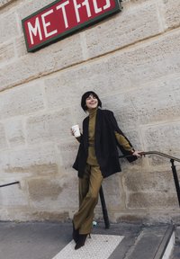 Femme en tenue vert olive et blazer noir tenant une tasse de café, appuyée sur une rambarde sous un panneau rouge "METRO" sur un mur de pierre.