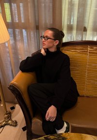 Pull noir à col roulé et pantalon large noir sur une personne assise dans un fauteuil en cuir marron avec un coussin texturé jaune. Lampe dorée à proximité.