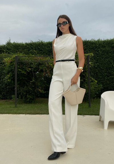 Mono blanco a medida con cuello alto y cinturón negro. Gafas de sol, bolso beige y zapatos negros completan el conjunto. Fondo verde.