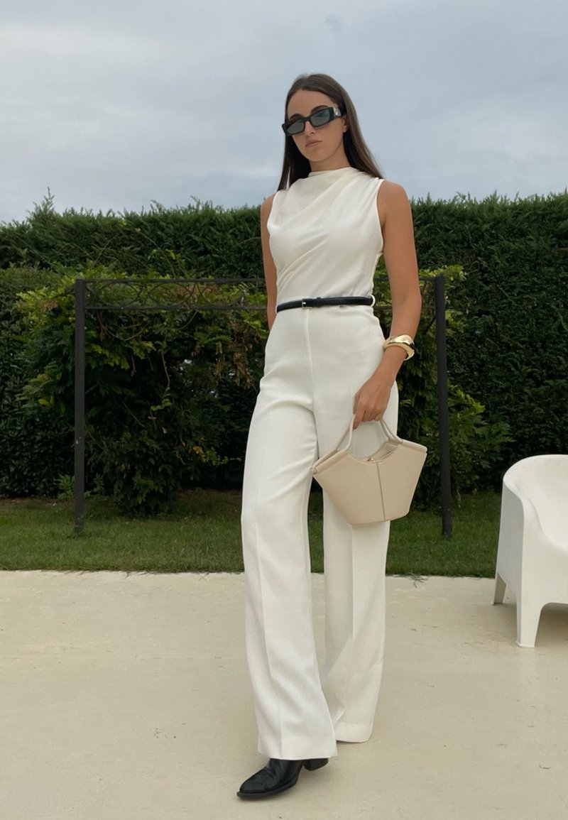 Weißer maßgeschneiderter Jumpsuit mit hohem Kragen und schwarzem Gürtel. Sonnenbrille, beigefarbene Handtasche und schwarze Schuhe runden das Outfit ab. Grüner Hintergrund.