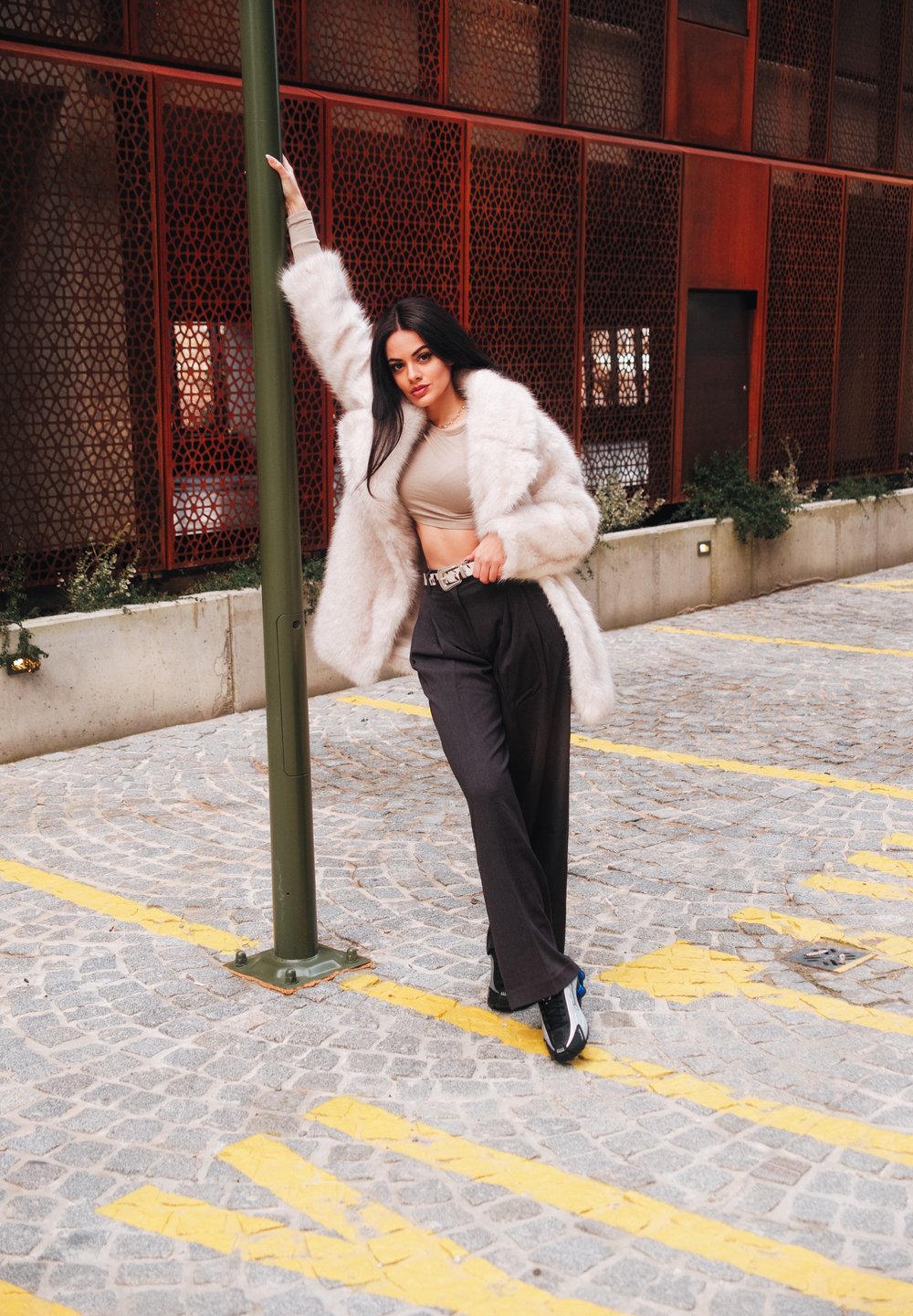 Faux fur jas in lichtbeige, getailleerde top, hooggevoerde zwarte broek en zwarte schoenen met blauwe accenten; model leunt tegen een groene paal.