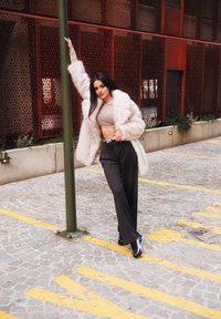 Faux fur jas in lichtbeige, getailleerde top, hooggevoerde zwarte broek en zwarte schoenen met blauwe accenten; model leunt tegen een groene paal.