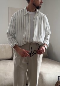 Homme en pantalon beige et chemise blanche à rayures tenant des lunettes noires, debout dans une pièce aux tons neutres avec un canapé beige en arrière-plan.