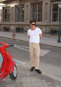 Balta T-krekls ar sarkaniem akcentiem, bēšas bikses un melni apavi, stāv blakus sarkanai skūterim uz pilsētas ielas ar akmens bruģi.