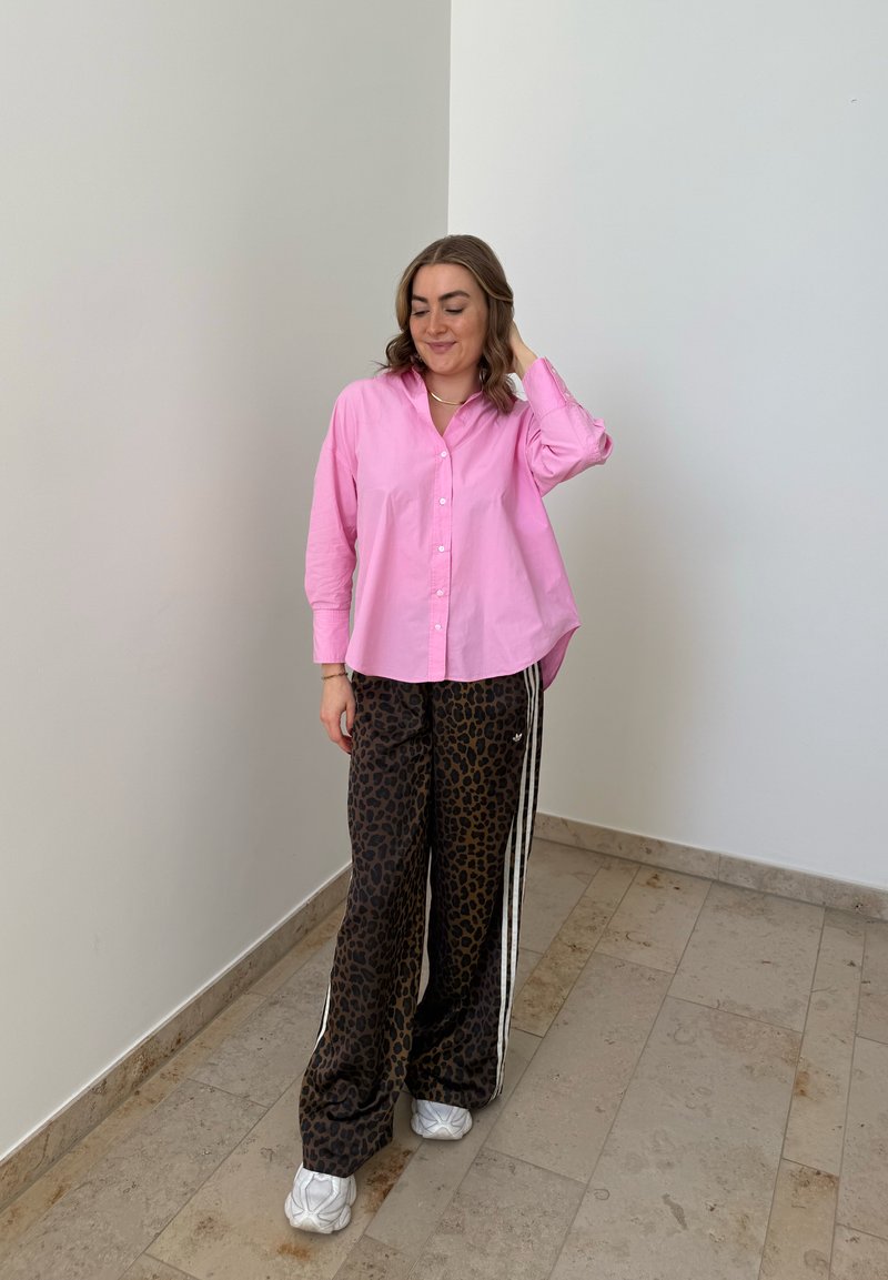 Donna in piedi all’interno che indossa una camicia rosa con bottoni, pantaloni larghi con stampa leopardata e strisce bianche, e scarpe da ginnastica bianche, toccandosi i capelli.