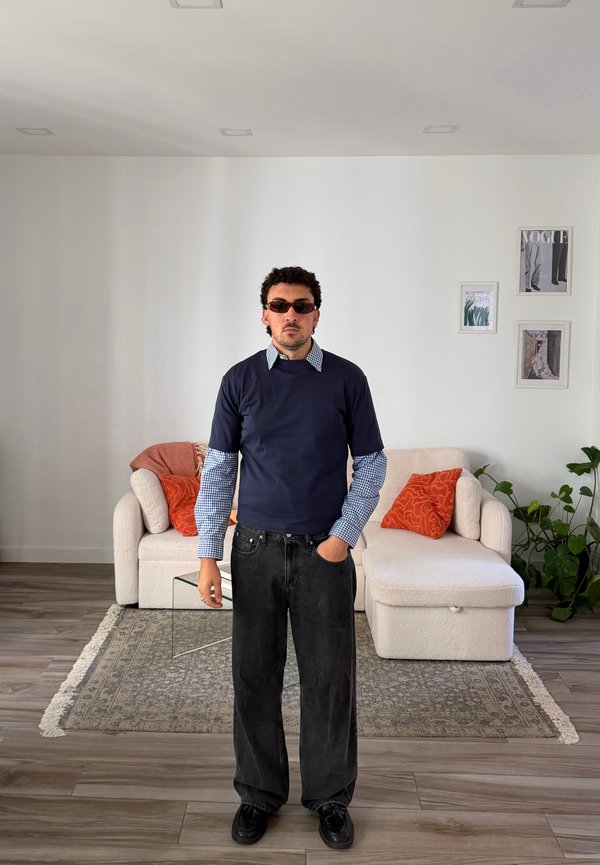 Mann mit lockigem Haar und Sonnenbrille steht im Wohnzimmer und trägt ein marineblaues Hemd über einem karierten Langarmhemd und schwarze weite Hose.
