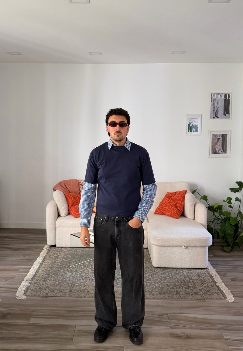 Homem com cabelo encaracolado e óculos de sol está na sala de estar usando camisa azul-marinho sobre mangas longas aos quadrados e calças largas pretas.