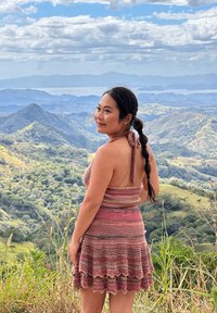 Femme aux cheveux tressés portant une robe en crochet rose debout sur une colline, regardant en arrière vers des montagnes verdoyantes sous un ciel bleu partiellement nuageux.