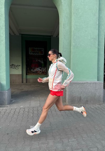 Corredor que lleva una chaqueta blanca ligera y transparente con detalles en verde azulado y pantalones cortos rojos. Zapatillas blancas con detalles en naranja y negro.