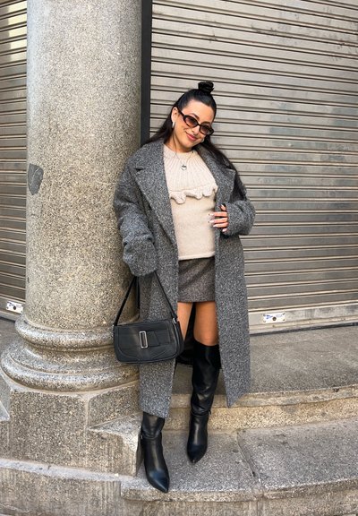 Abrigo gris de gran tamaño sobre un suéter beige con volantes, falda de cuadros y botas negras hasta la rodilla. Un bolso negro completa el conjunto.