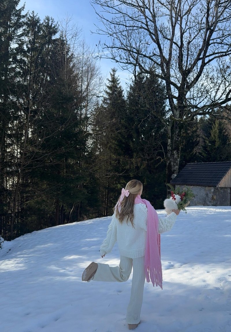 Pull et pantalon en maille épaisse blanche, associés à une écharpe rose clair à franges. Tenant un bouquet de fleurs, debout sur un sol recouvert de neige.