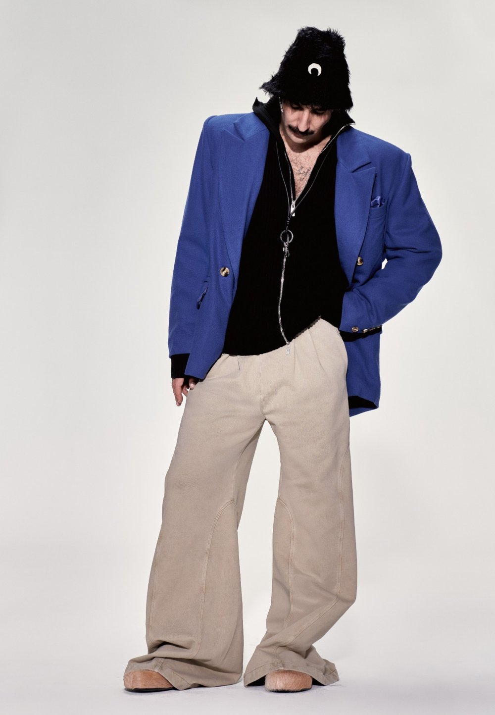 Overdimensioneret blå blazer, sort lynlås-sweater, lysebeige vidde bukser og en sort fuzzy hat med en detaljeret halvmåne.