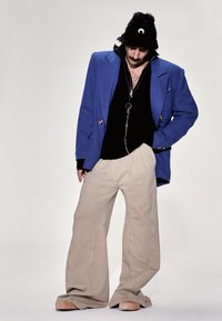 Overdimensioneret blå blazer, sort lynlås-sweater, lysebeige vidde bukser og en sort fuzzy hat med en detaljeret halvmåne.