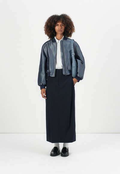 Chaqueta bomber de mezclilla con cuello y puños acanalados, falda azul marino, mocasines negros y un bolso de hombro azul, sobre un fondo blanco liso.