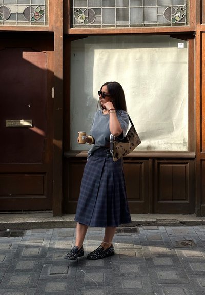 Camiseta gris, falda de cuadros hasta la rodilla con hebilla lateral, zapatos negros con tachuelas y bolso de mano de estampado de leopardo. Sosteniendo una taza de café.