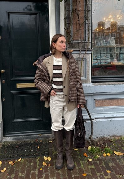 Mujer con ropa de otoño en capas que incluye chaqueta acolchada a cuadros, suéter a rayas, pantalones beige y botas altas marrones, de pie junto a una puerta y ventana negras.