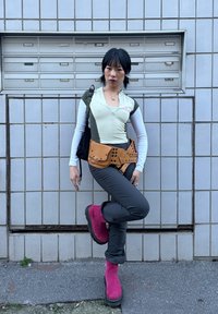 Jeune femme posant avec une jambe levée, portant un haut vert et blanc superposé, un pantalon gris, des bottes roses, un sac ceinture marron et un sac à bandoulière noir, devant des boîtes aux lettres.