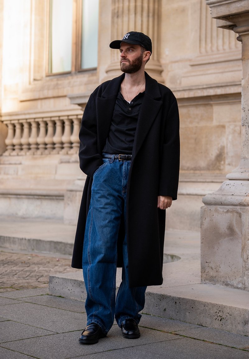 Homme barbu portant un manteau noir, une chemise noire, un jean bleu et des baskets noires, debout sur un pavé en pierre près d'un bâtiment classique.