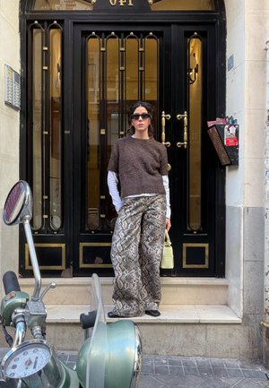 Mujer con gafas de sol, suéter café, pantalones con estampado de serpiente y sosteniendo un bolso amarillo, de pie frente a una puerta negra con detalles dorados. Hay un scooter cerca.