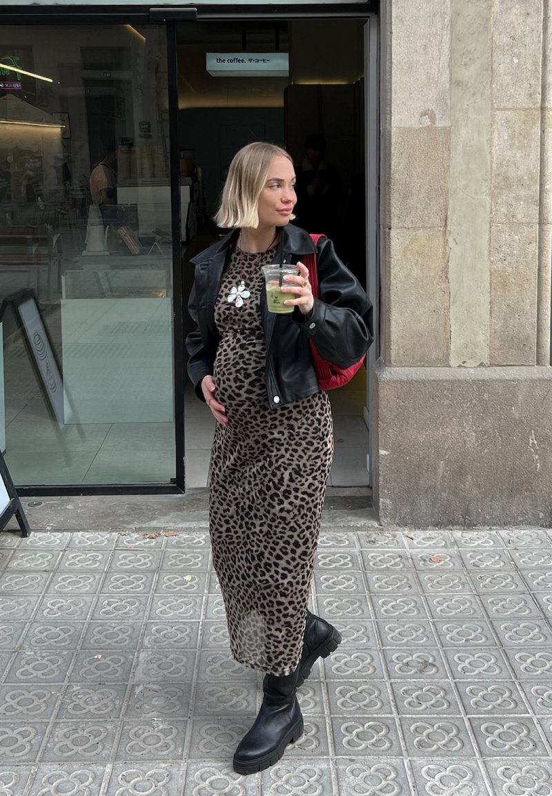 Μαύρη δερμάτινη τζάκετ πάνω από φόρεμα με λεοπάρ σχέδιο, μαύρες μποτάκια μέχρι τον αστράγαλο, κρατώντας ένα ποτό, στέκεται σε κυβόλιθους κοντά στην είσοδο ενός καφέ.