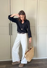 Zwarte linnen overhemd met knopen, witte wijde broek, bruine riem, grijze slippers en een geweven stro tote bag met leren accenten.
