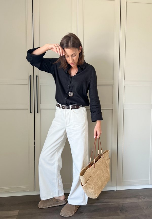 Zwarte linnen overhemd met knopen, witte wijde broek, bruine riem, grijze slippers en een geweven stro tote bag met leren accenten.