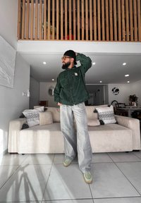 Grüne Cordjacke mit Knopfverschluss, graue weite Hosen und hellgrüne Schuhe in einem modernen Wohnraum mit einem strukturierten Sofa.
