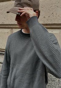 Ung man som justerar skärmen på en brun keps, klädd i en gråtexturerad tröja, stående mot en beige stenvägg.