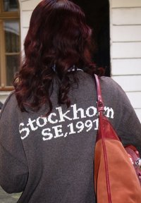 Grå striktrøje med hvid skrift "Stockholm SE. 1991" på ryggen; glat tekstur. Rød håndtaske med et kontrastfarvet panel.