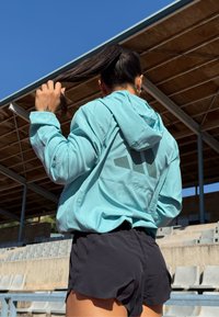 Helle blaue Kapuzenjacke mit einem Logo auf dem Rücken, kombiniert mit schwarzen Shorts. Die Jacke hat einen lässigen Schnitt und lange Ärmel.