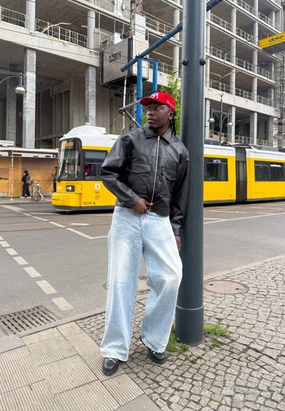 Chaqueta de cuero negra con dos bolsillos frontales; jeans de pierna ancha color azul claro; gorra roja. Entorno urbano con un tranvía amarillo al fondo.